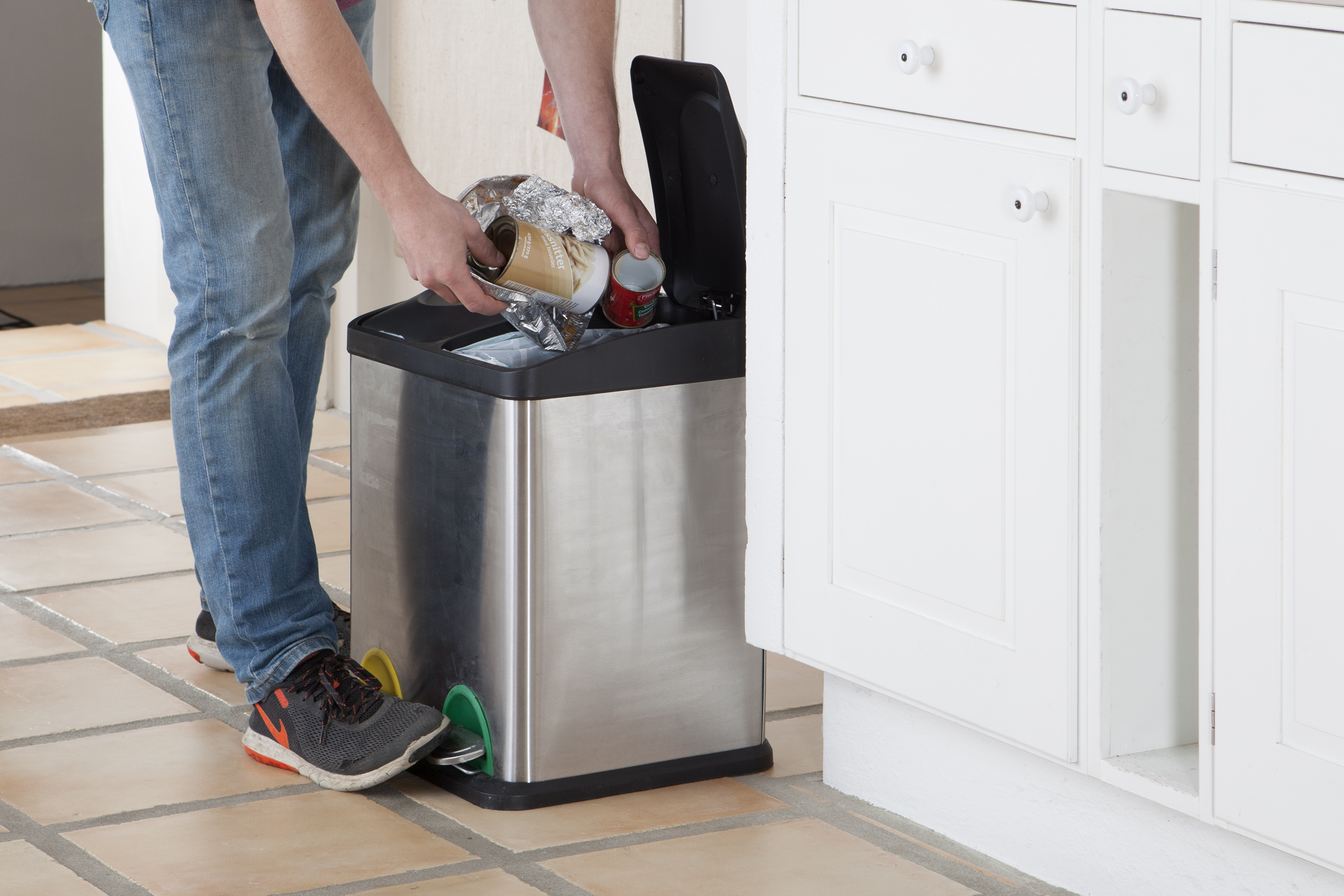 Person i jeans og sneakers smider affald i en skraldespand af rustfrit stål i et køkken med hvide skabe og flisegulv.